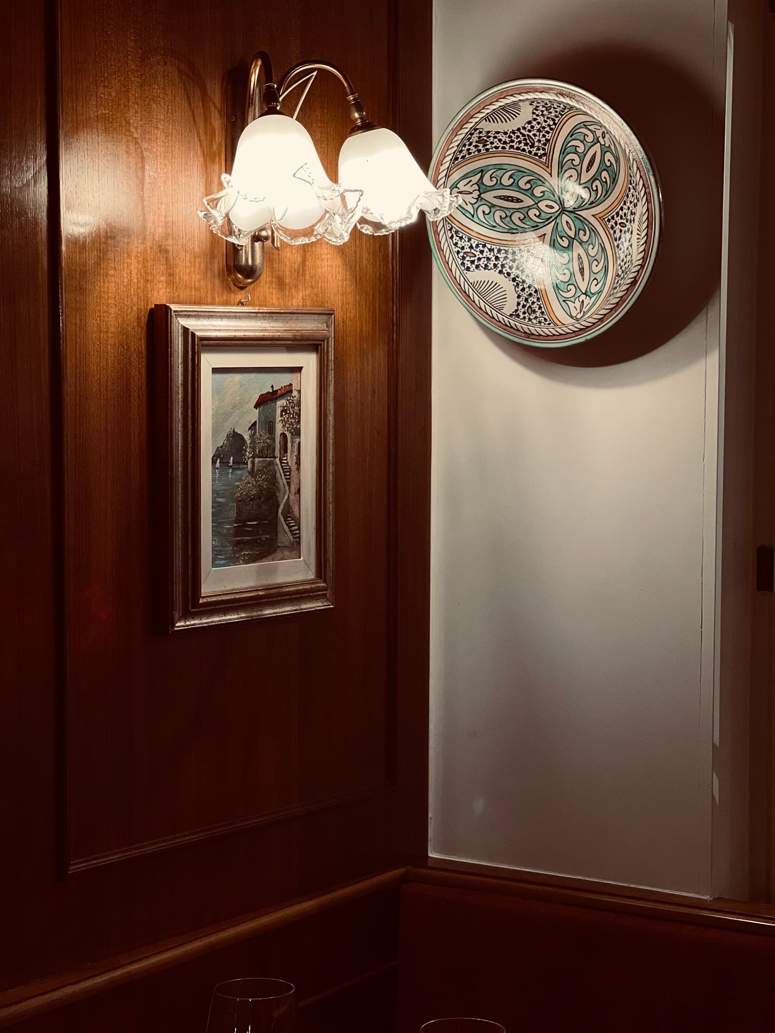 Une applique, une petite peinture de village italien au bord de l’eau et une assiette en céramique peinte à la main dans la salle boisée de la trattoria.
